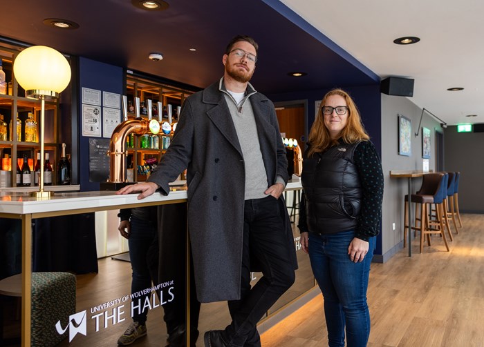 Two people stand in a modern bar area with the sign "The Halls, University of Wolverhampton" visible on the counter. Bottles and taps are displayed in the background.