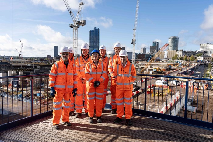 Group of seven people in orange construction overalls on balcony overlooking the large Hs2 building site in Birmingham city centre