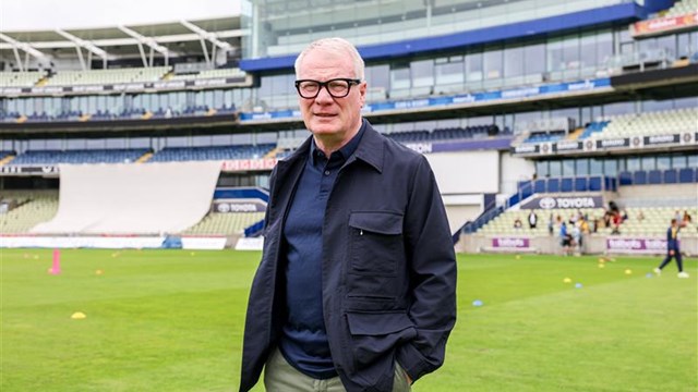 A man wearing glasses and a dark jacket stands on a sports field with a stadium and spectator stands and seating in the background.