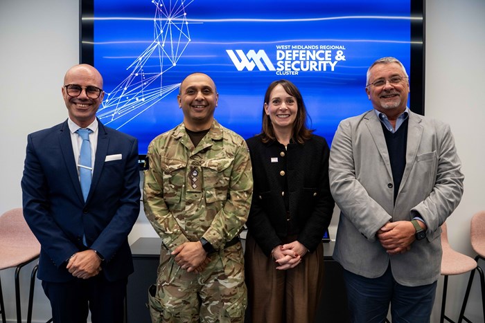 Four people stand smiling in front of a "Defence & Security" display. From left: a man in a suit, a person in military attire, a woman in a black jacket, and a man in a grey blazer.