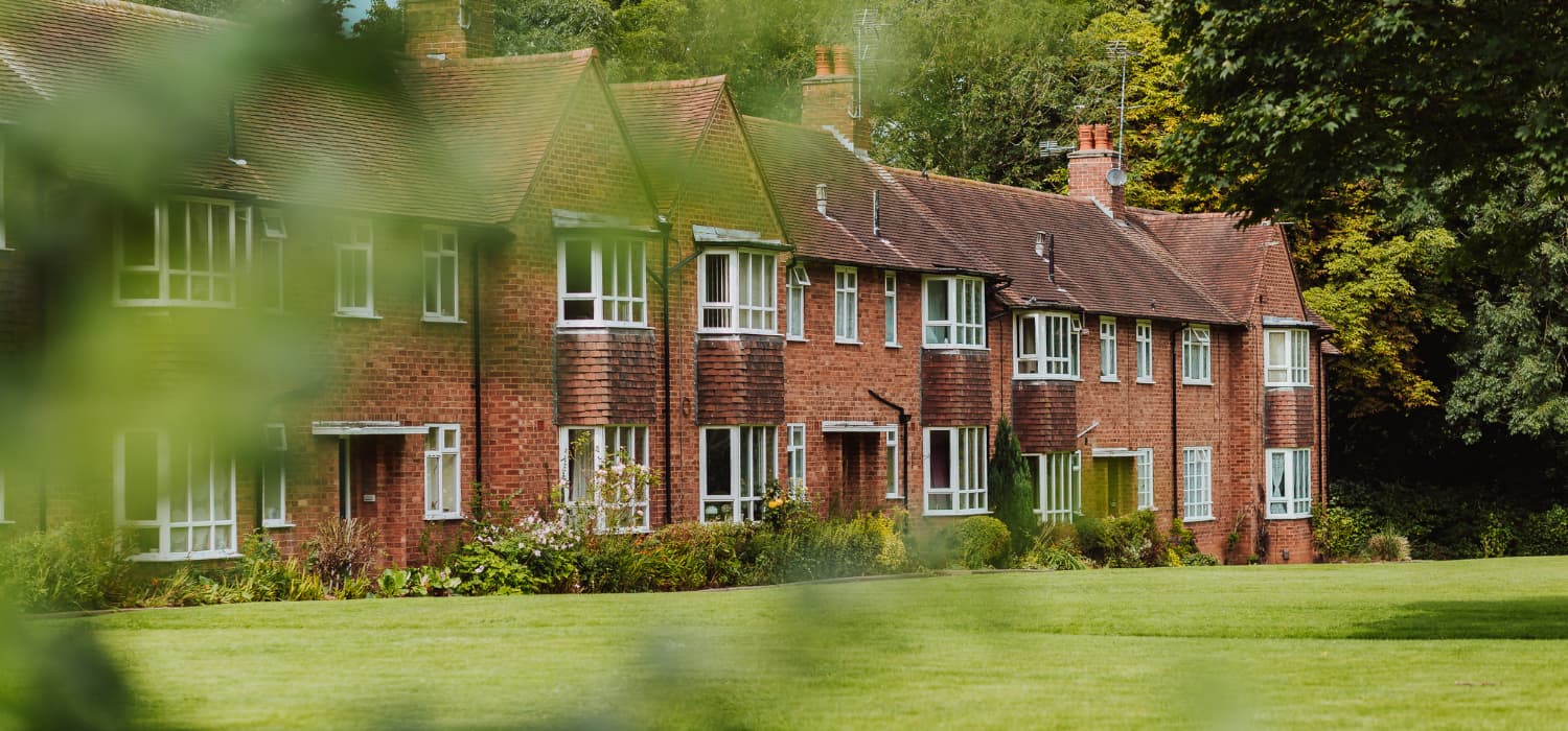 Row of red‑brick houses with large windows, viewed across a green lawn and partially framed by foliage