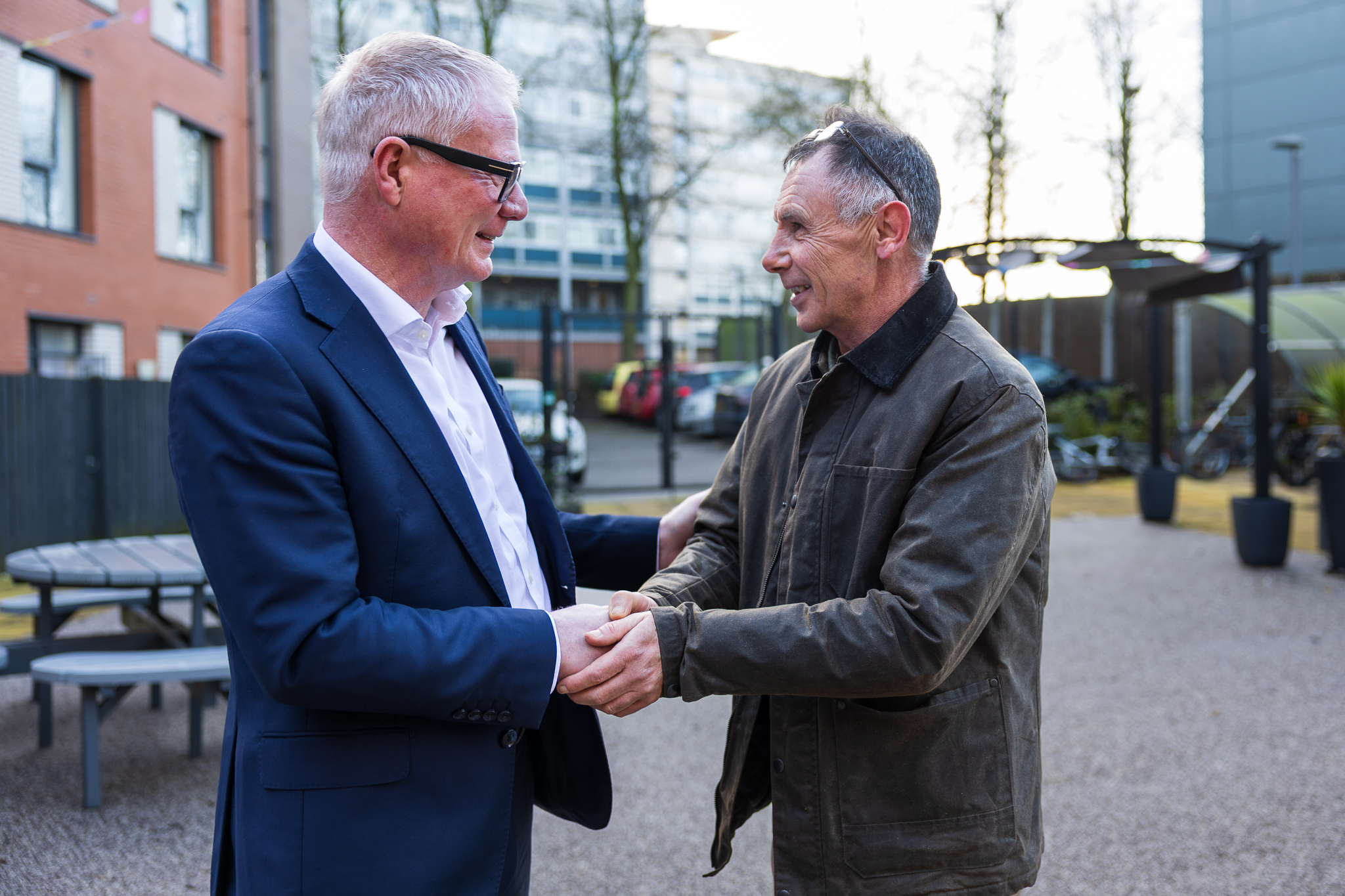 Two men smiling and shaking hands outdoors, one in a suit and the other in a casual jacket. The setting is an urban area with a relaxed, cordial look.
