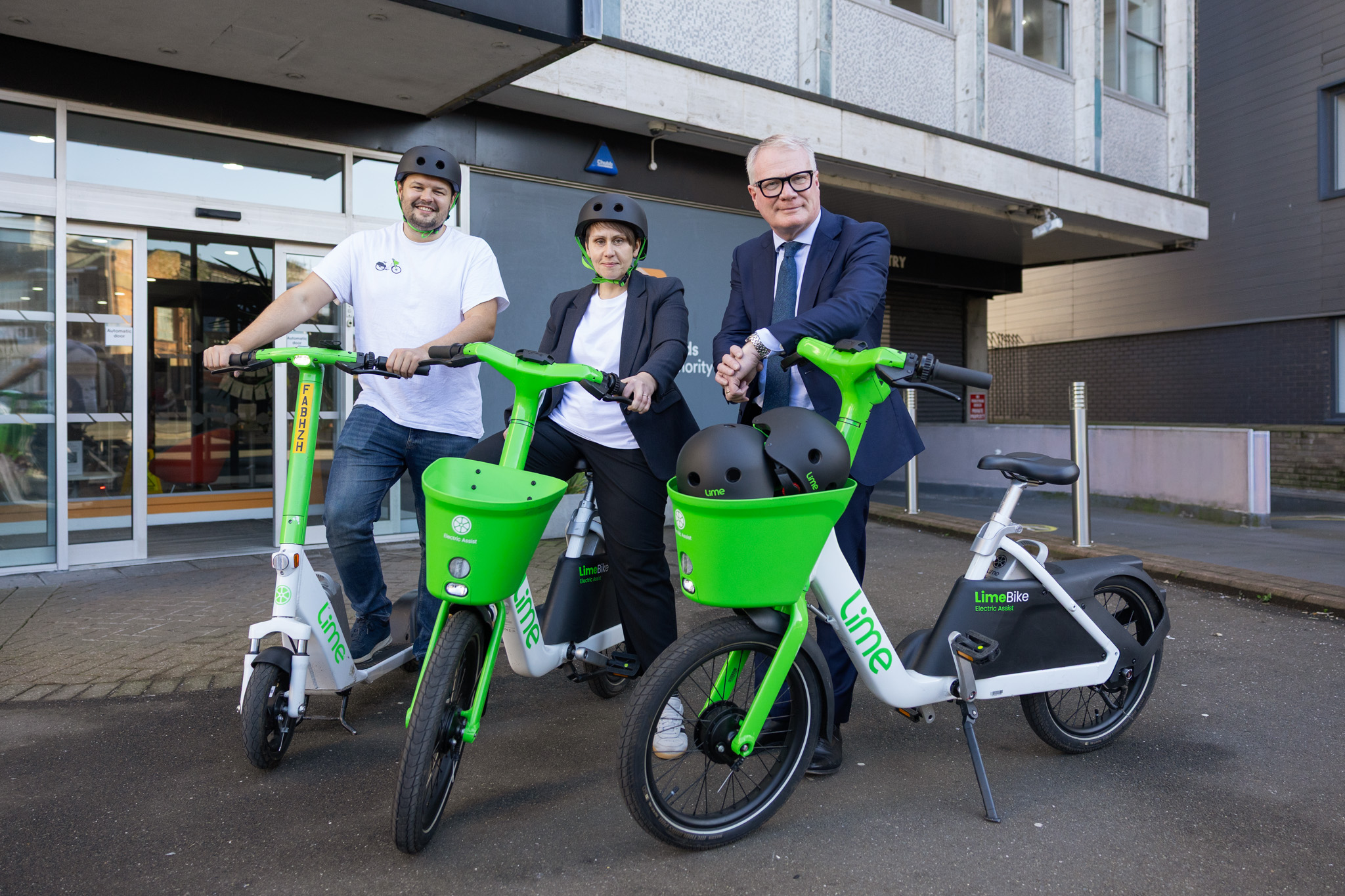 Three people standing behind the handlebars of an e-scooter and two green and white coloured e-bikes