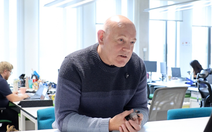 Bald man in a dark sweater leans forward over an office desk, hands clasped, speaking seriously in a bright, busy open-plan workspace.