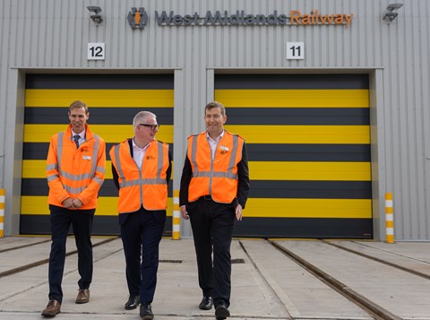 Three men, in orange hi-vis vests walking in front of large metal warehouse building which fills rest of the picture. There are two black and yellow striped garage doors and a sign above says West Midlands Railway