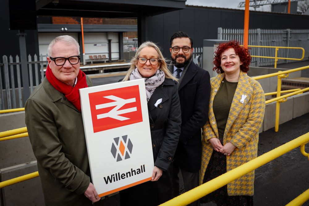 Group of four people holiding a white sign with railway insignia and Willenhall written. Behind them is the entrance to the station with a paltform and bench visibl