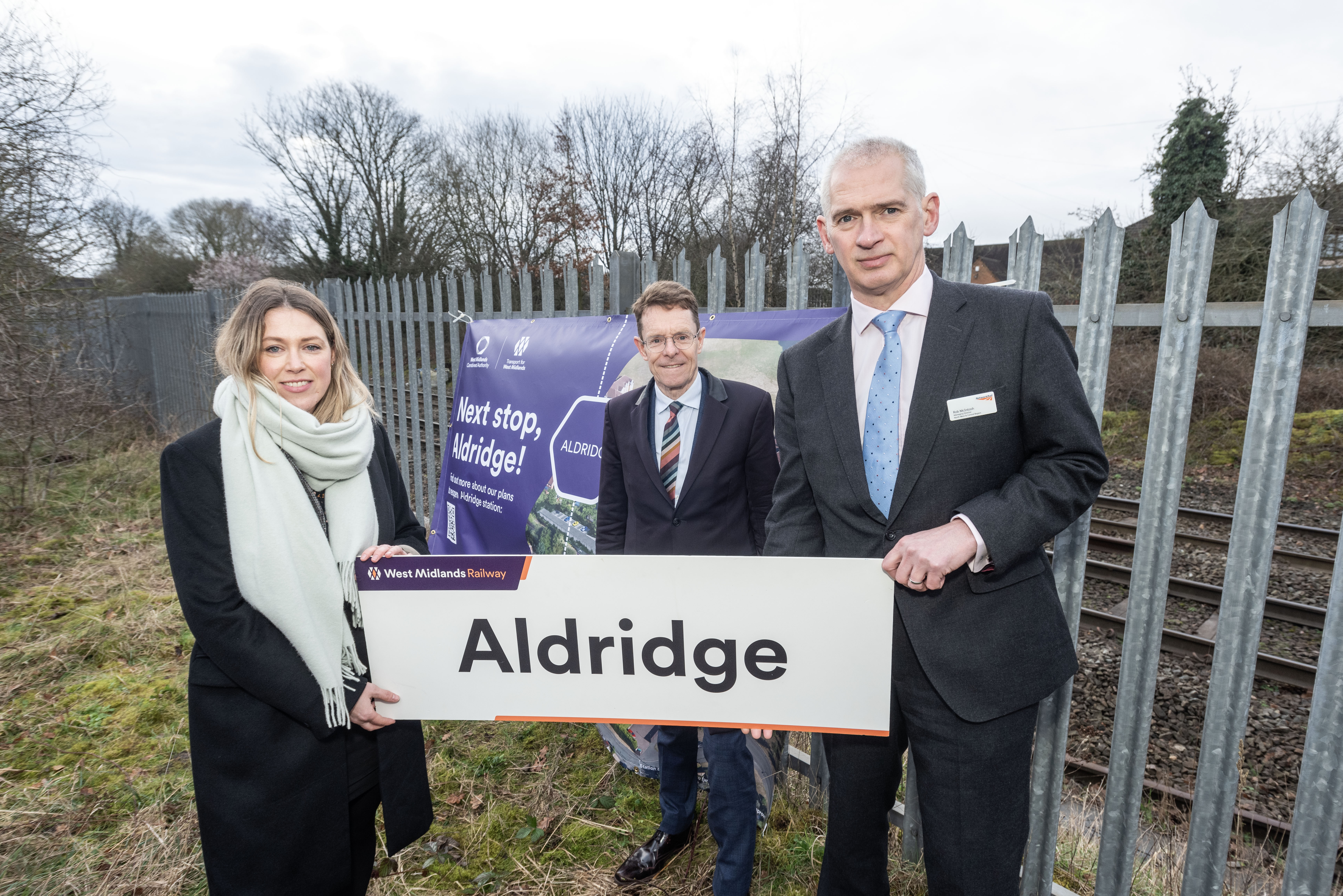 Kate Trevorrow, TfWM rail delivery director, Andy Street Mayor of the West Midlands and Rob McIntosh, managing director for Network Rail's North West and Central Region holding an Aldridge Station sign with the rail line behind a fence in the background