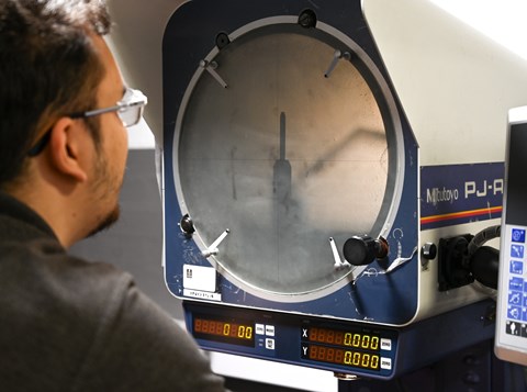 A person observes a precision measurement on a Mitutoyo profile projector with numeric digital displays and a control panel.