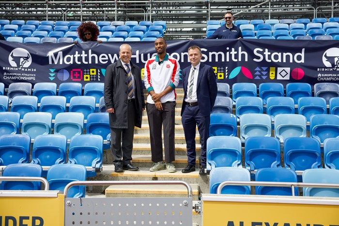 Three individuals stand on stadium steps with a "Matthew Hudson-Smith Stand" banner. They are surrounded by blue seats