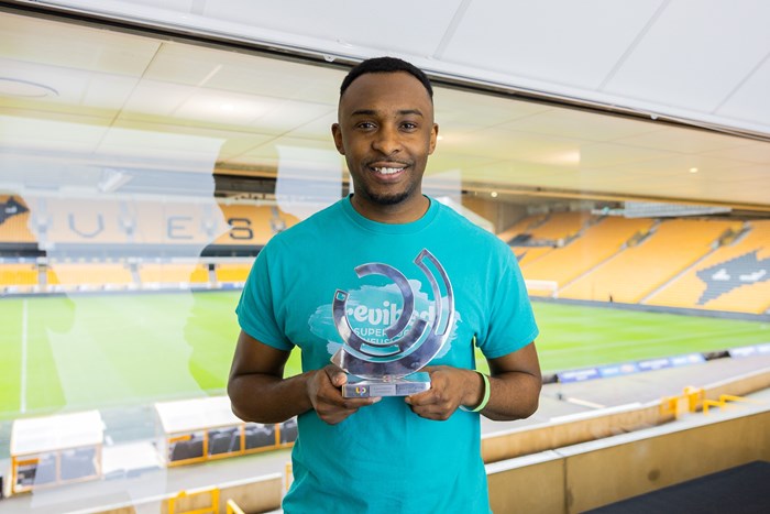 A smiling man in a turquoise T-shirt holds a transparent award with a stadium visible in the background. The scene conveys a sense of achievement.