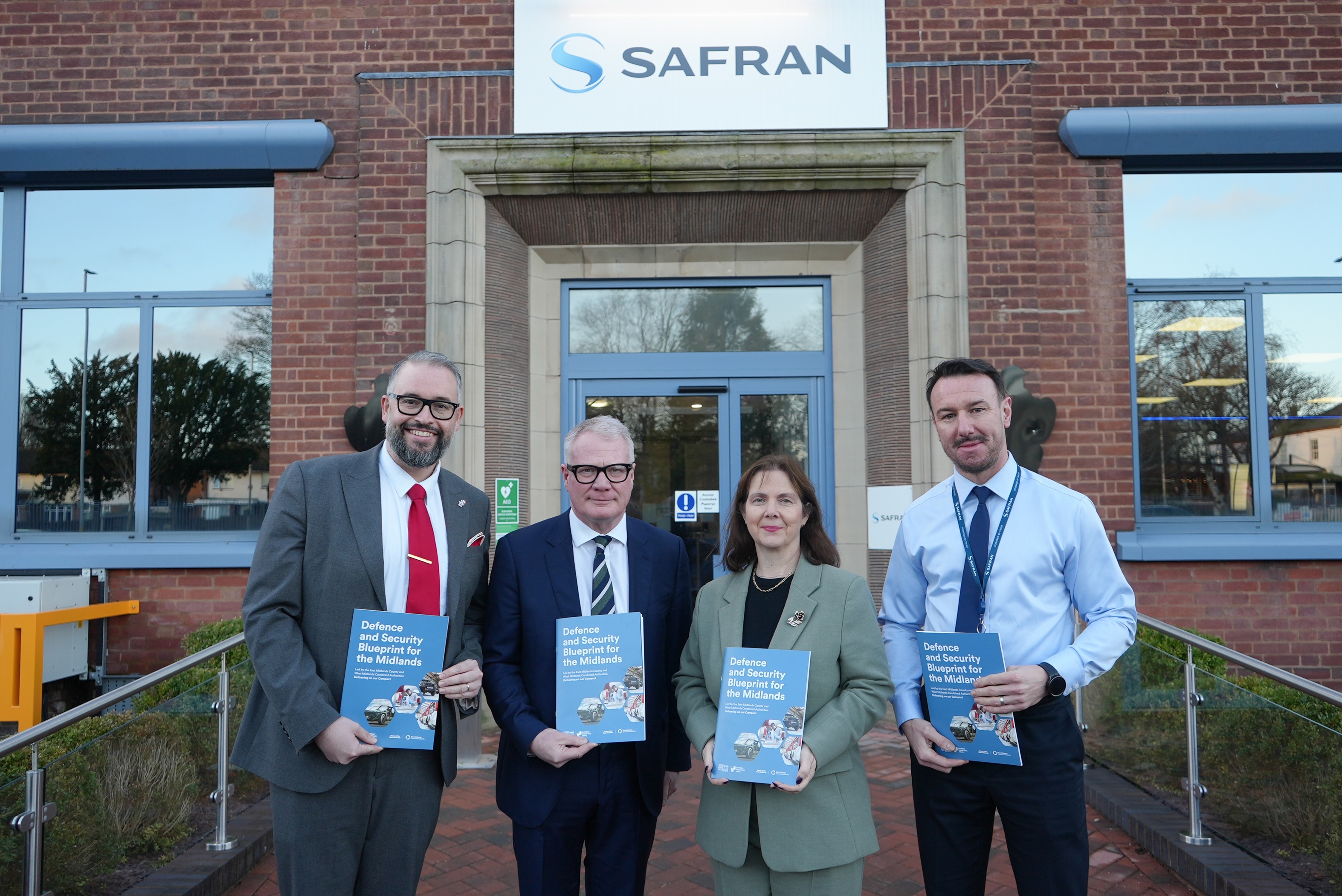 Four individuals stand smiling in front of a building with a "SAFRAN" sign, holding blue books. The mood is professional and positive.