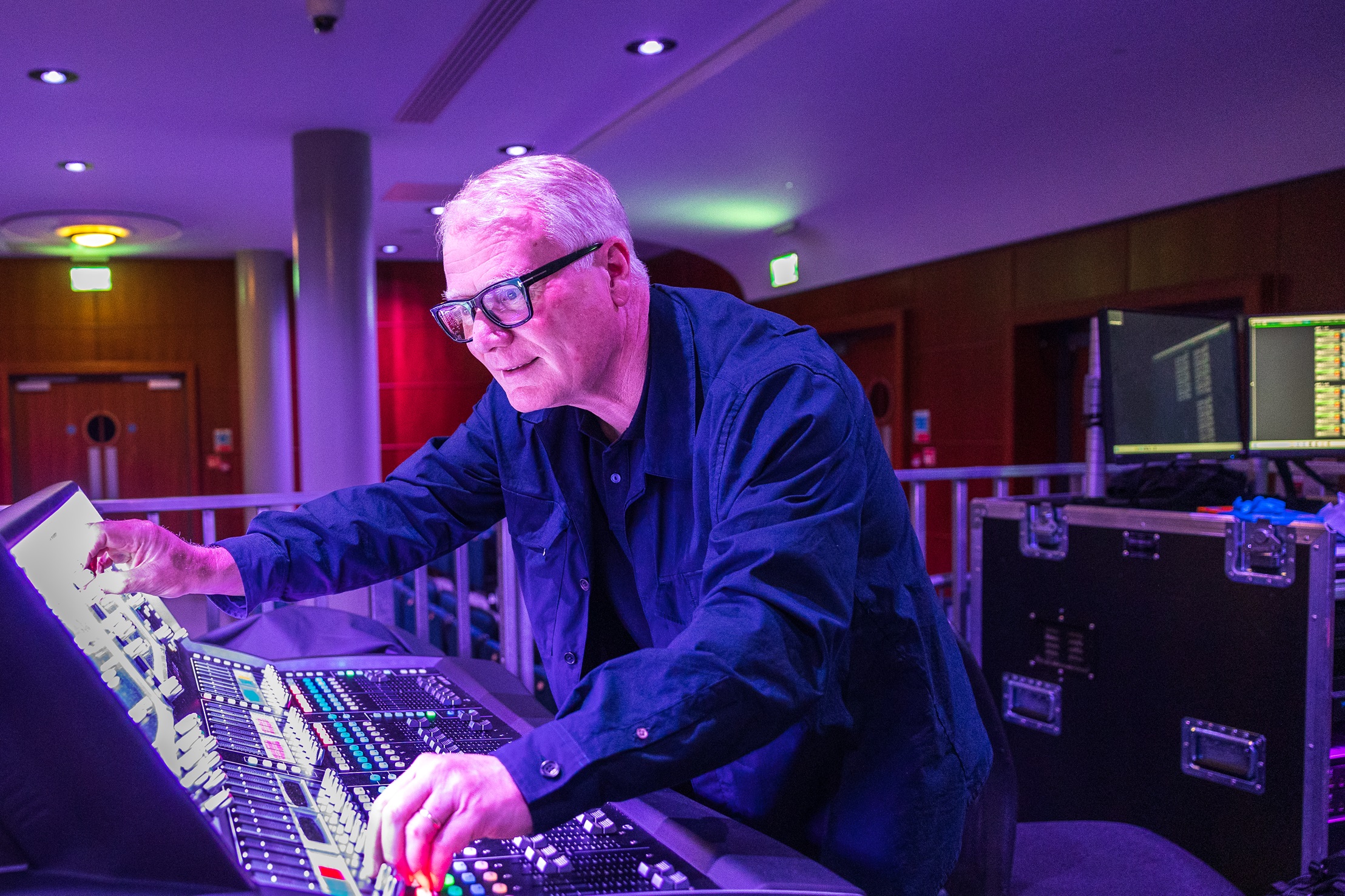 A man with glasses wearing a blue shirt operates a professional audio mixing console in a dimly lit room with purple ambient lighting and monitors.