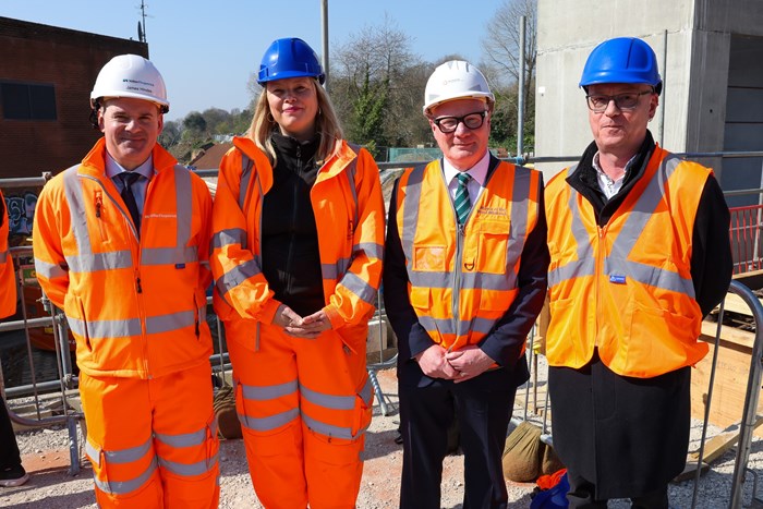 Four people in hard hats and hi-vis oragne outfits in front of scaffolding and a concrete pillar