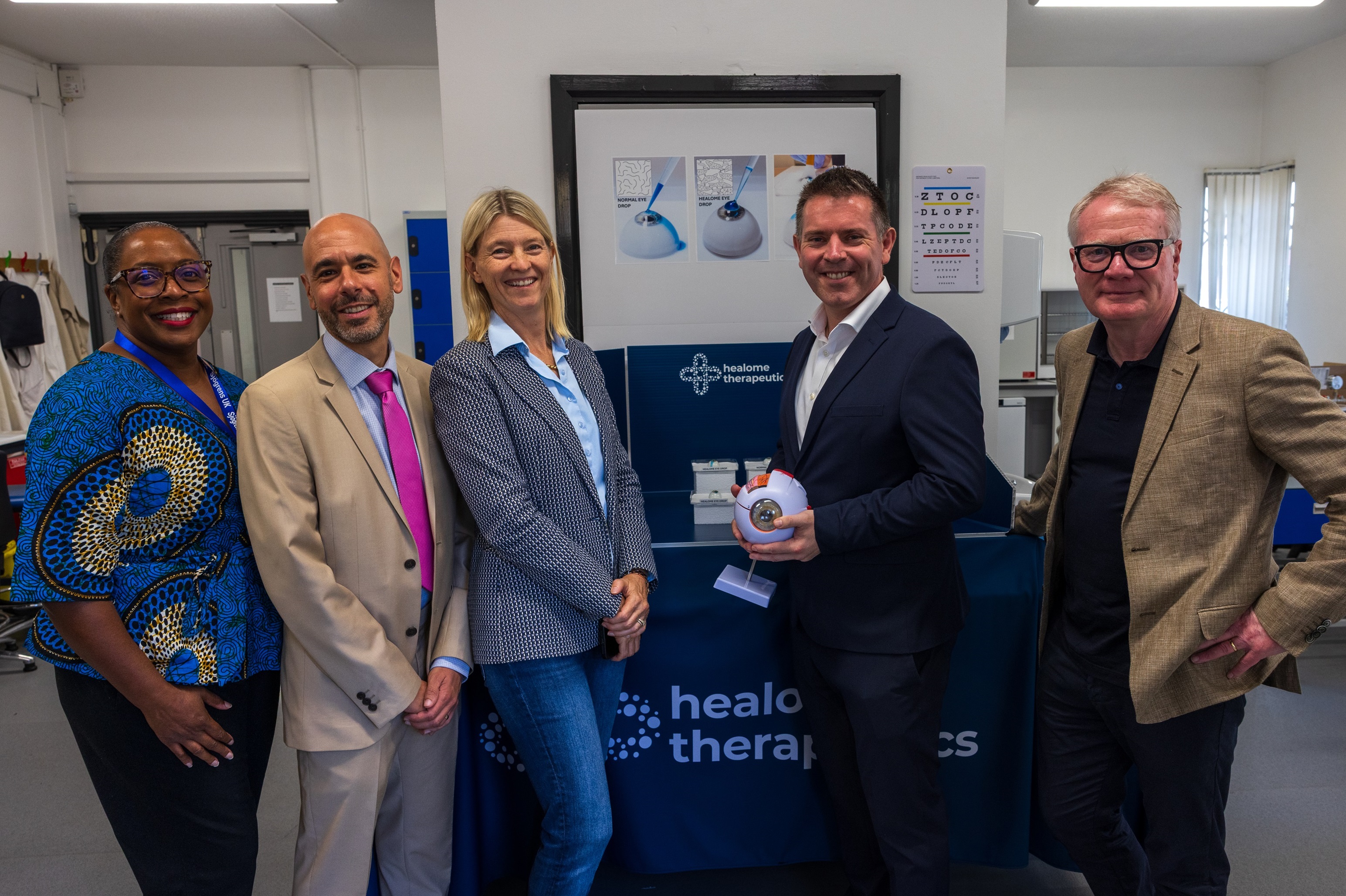 Five professionals pose at a healthcare therapy event; a suited man holds a small device beside a branded table, smiling for the camera.