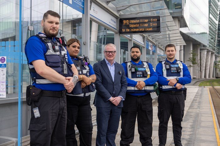 A group of five people stood under a tram stop canopy with a city centre office behind in the distance. Four of the line up are wearing Transport Safety uniforms, including bright blue polo shirts with a dark stab-proof vest over the top. In the centre of the line is Mayor Richard Parker wearing his suit jacket.