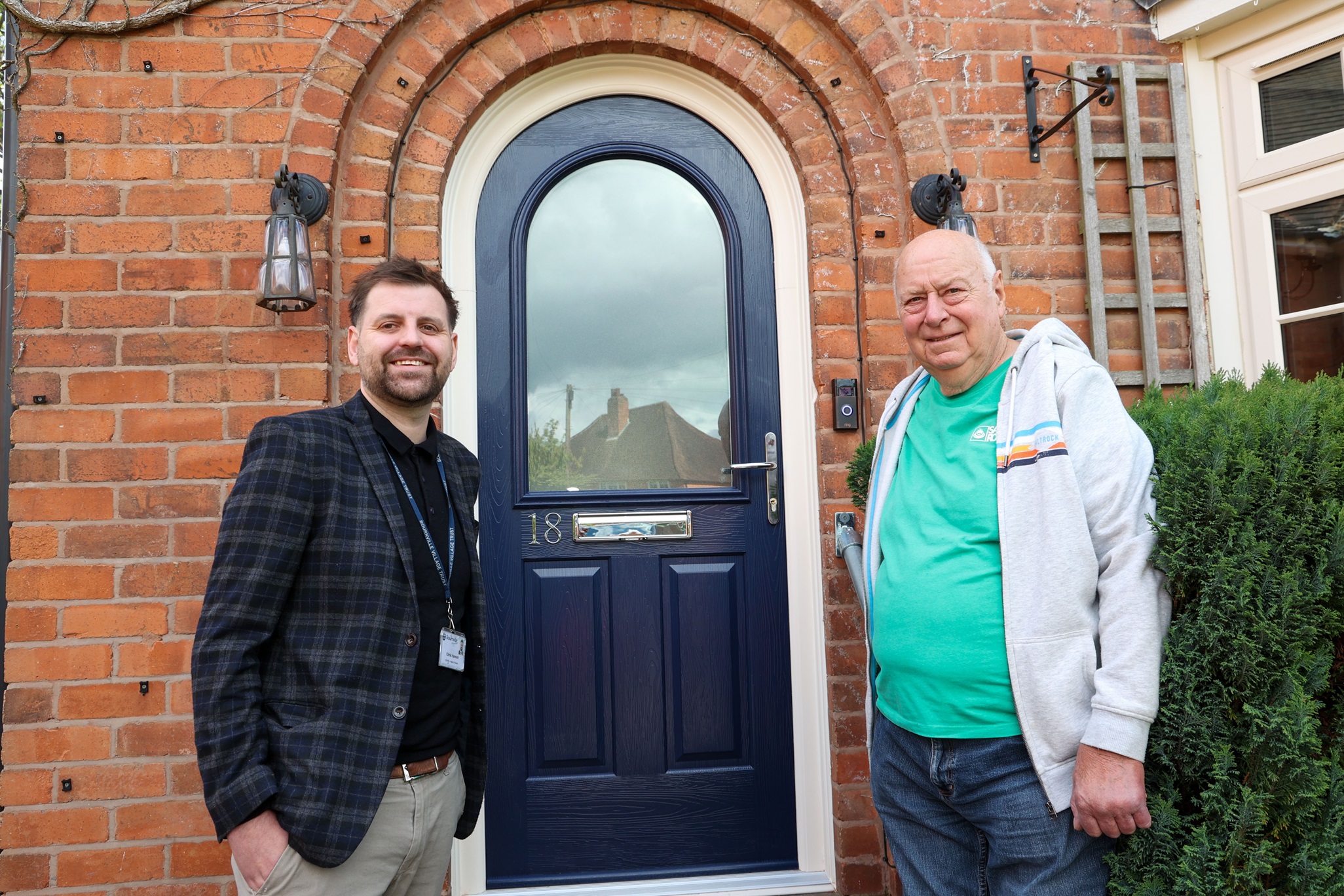 Two men smile in front of a brick house with an arched blue door. One wears a checked jacket, the other a teal shirt and gray hoodie. 