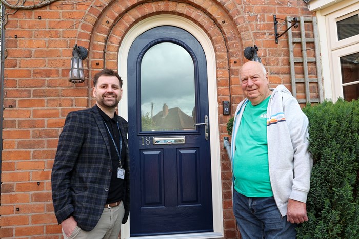 Two men smile in front of a brick house with an arched blue door. One wears a checked jacket, the other a teal shirt and gray hoodie.