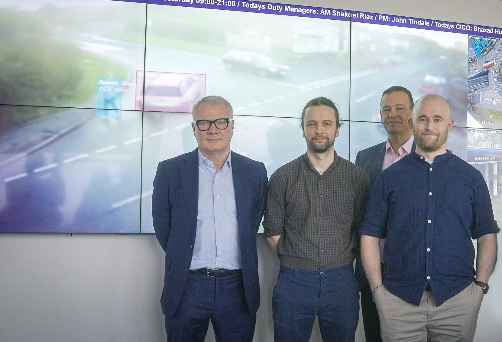 Four men standing in front of video screen showing image of cars and pedestrian at a jucntion