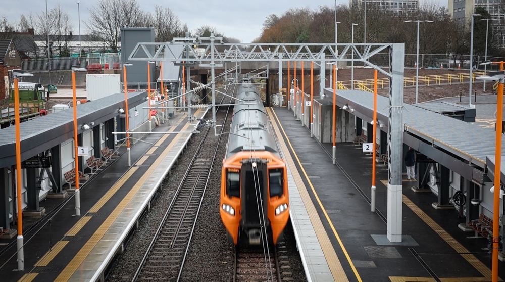 Aerial view from bridge over railway line showing train with yellow front passing through a station. Platforms and shelters on either side with organe pole lampposts and signs.