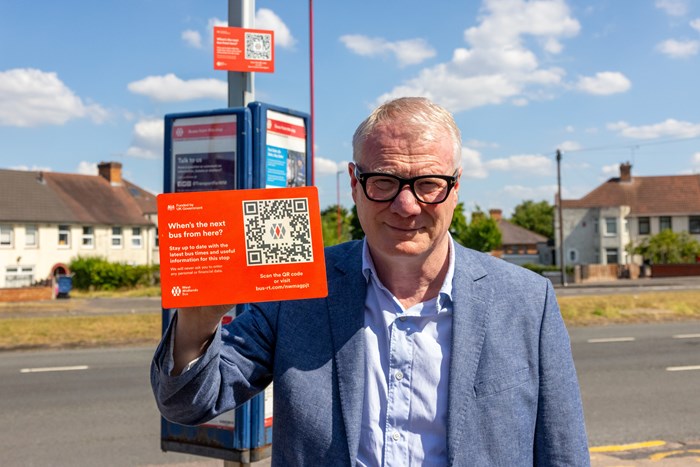 Mayor holding a QR sign, he's on the roadside in front of a bus stop pole with one of the codes above the timetable.
