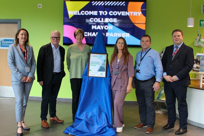 Six adults pose beside a framed certificate on a blue-draped stand in Coventry College’s bright lobby, beneath a welcome screen; the mood is formal and celebratory.