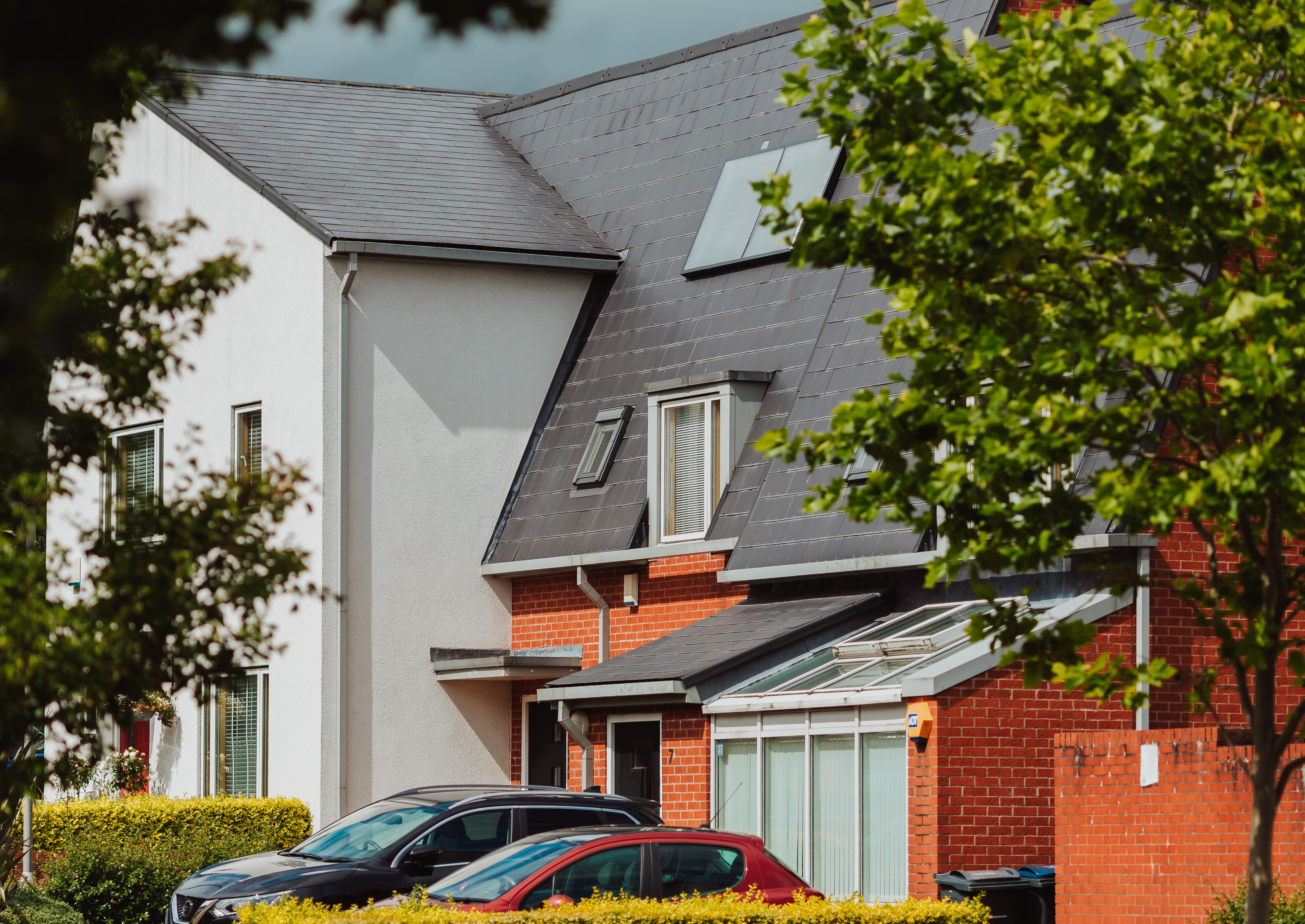 Modern semi-detached houses with sloped roofs, red brick and white walls, two parked cars in front, and green trees partially framing the view