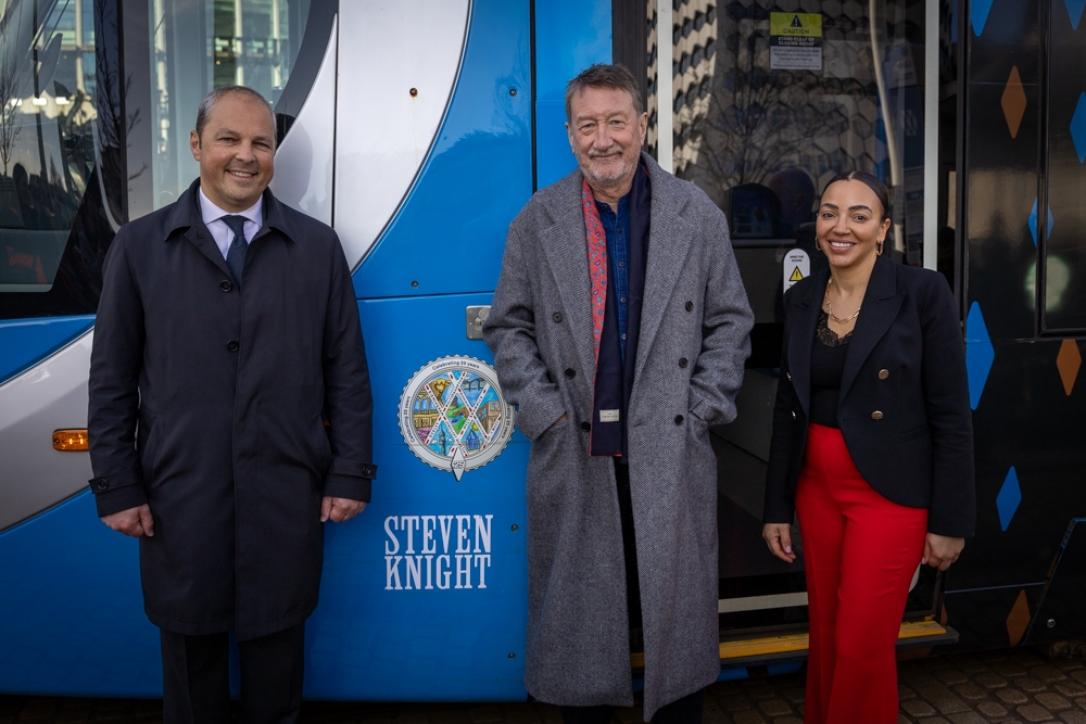 Three people lined up alongside a tram with the name Steven Knight on the side