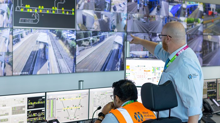 And operator sits at a desk in front of a wall of monitor screens with tram line views. Another operator stands at his side pointing.