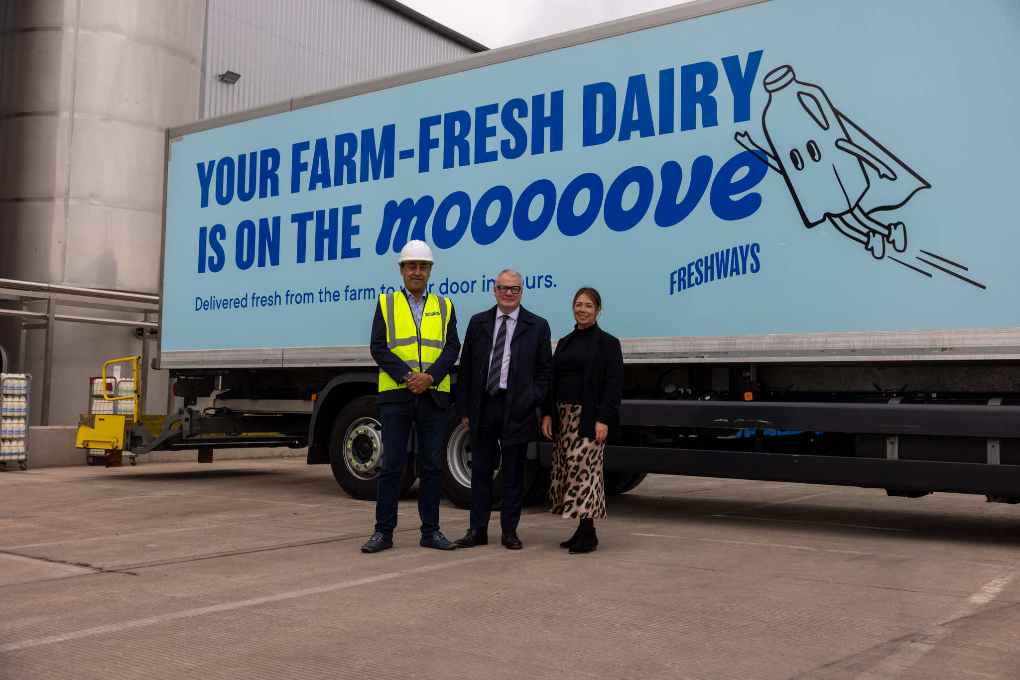 Three people standing in front of a blue lorry - the side of which bears a light blue design - with words 'your farm fresh dairy is on the moooove'. Behind that is a factory building and to the side part of steel piping and silo