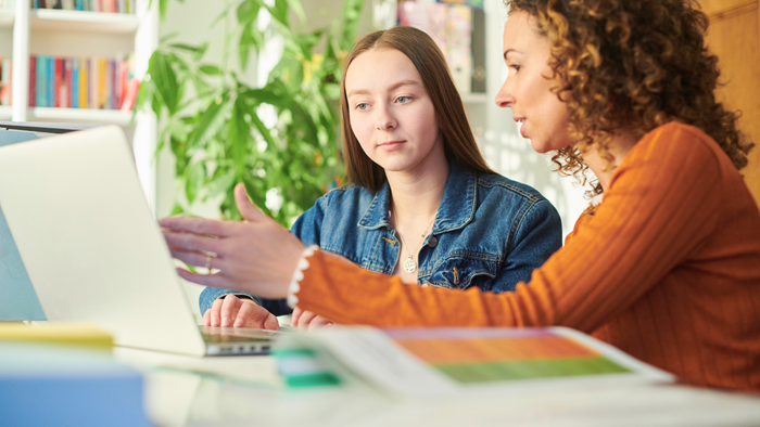 A woman and a girl sat in front of a laptop computer. The woman looks like she is giving advice while the girl listens.