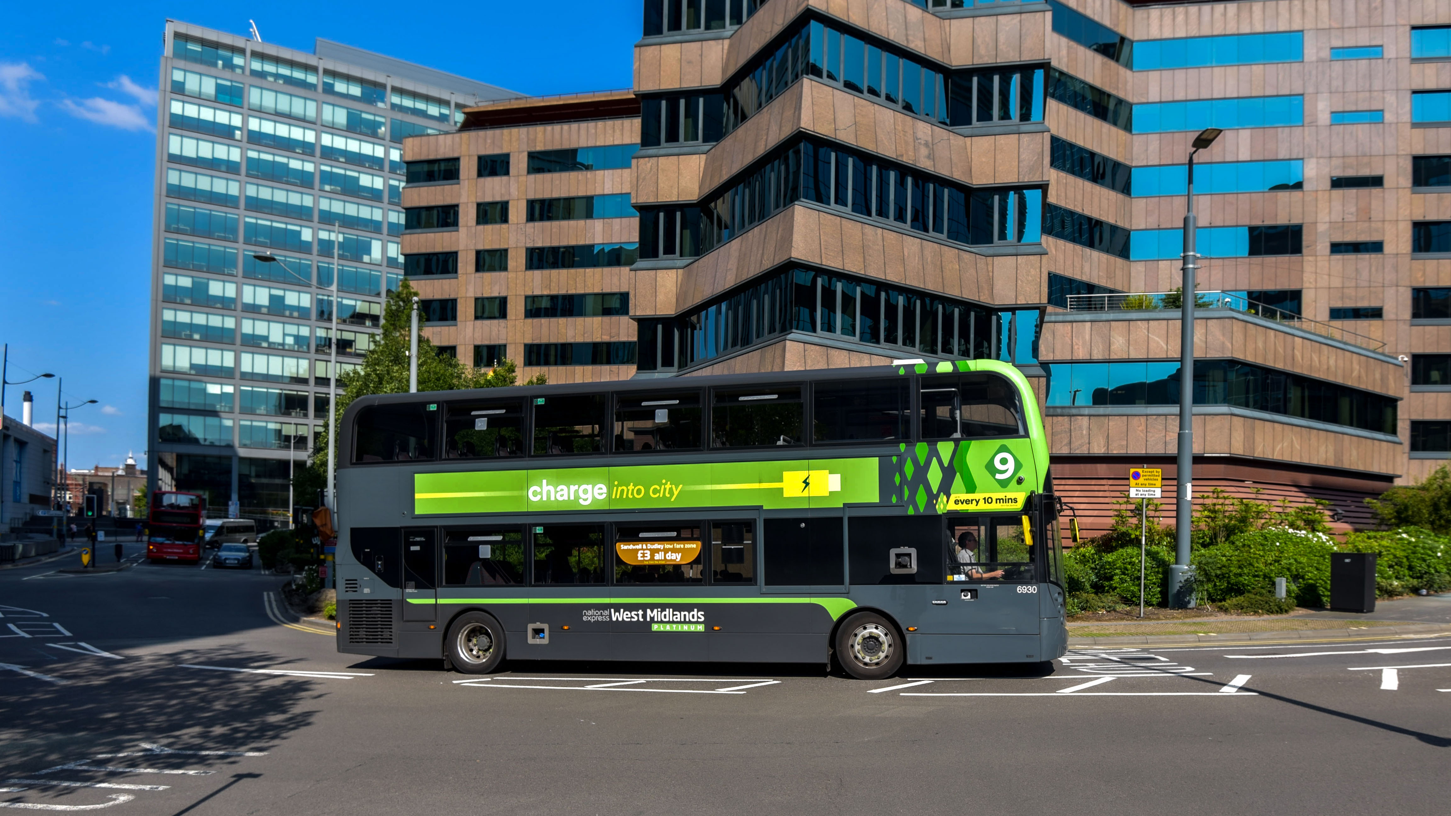 A grey and bright green electric double-decker bus operated by National Express West Midlands travelling through a city intersection. The bus features green branding along its side with the slogan "charge into city" and a battery icon, set against a backdrop of multi-story glass and brown office buildings under a clear blue sky.