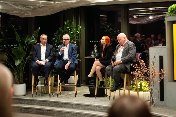 Four speakers seated on a stage during a panel discussion at an indoor business event, with microphones, audience members, and plants visible around the venue.