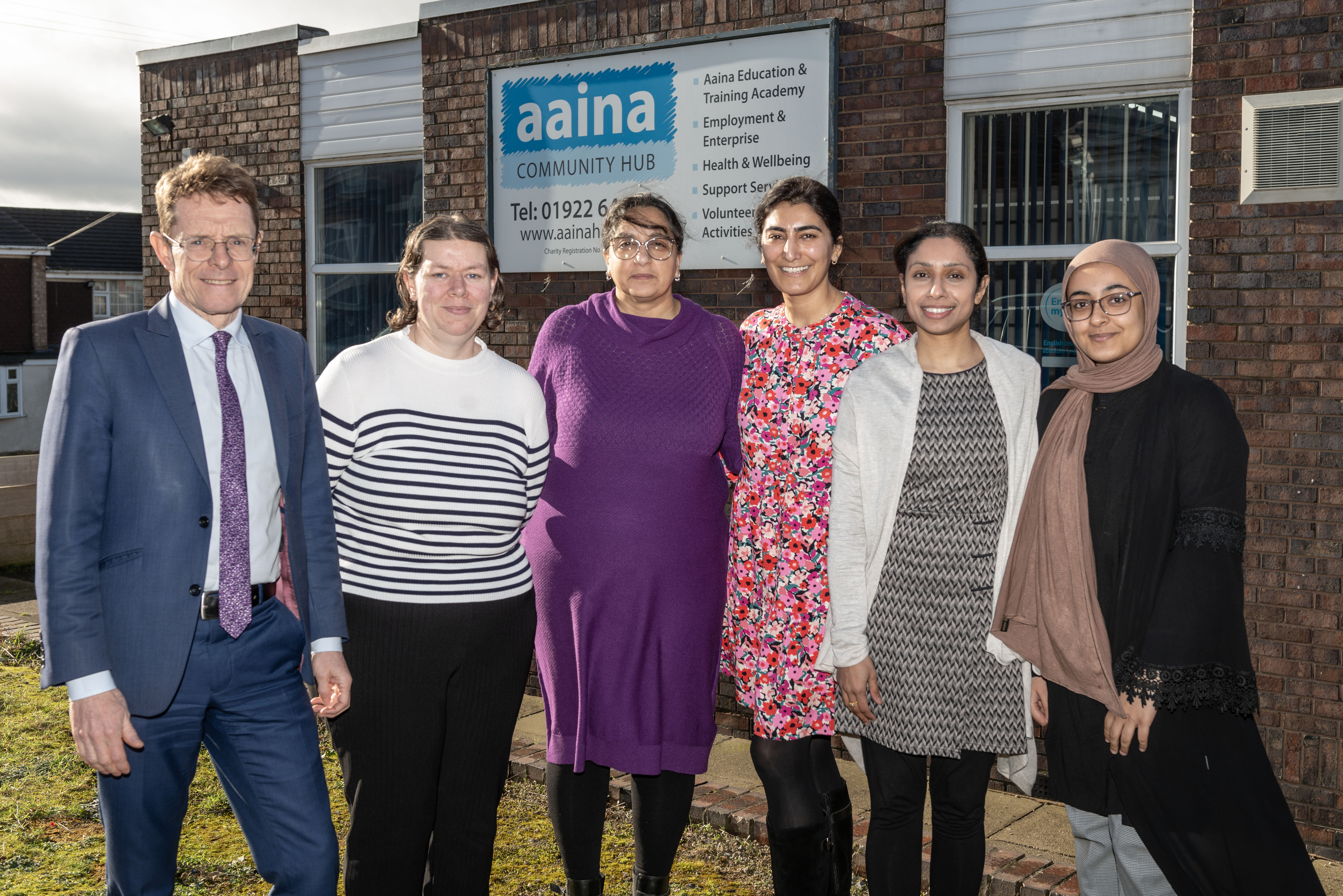 With the Aaina Hub in the background, Andy Street, Mayor of the West Midlands and WMCA chair, Carolyn Dews, A'isha Khan, Aaina Community Hub chief executive, Sharonjit Clare, independent chair of the West Midlands Race Equalities Taskforce, Sharon Khakh and Mahaam Arooj are pictured in the foreground standing in a line and smiling at the camera