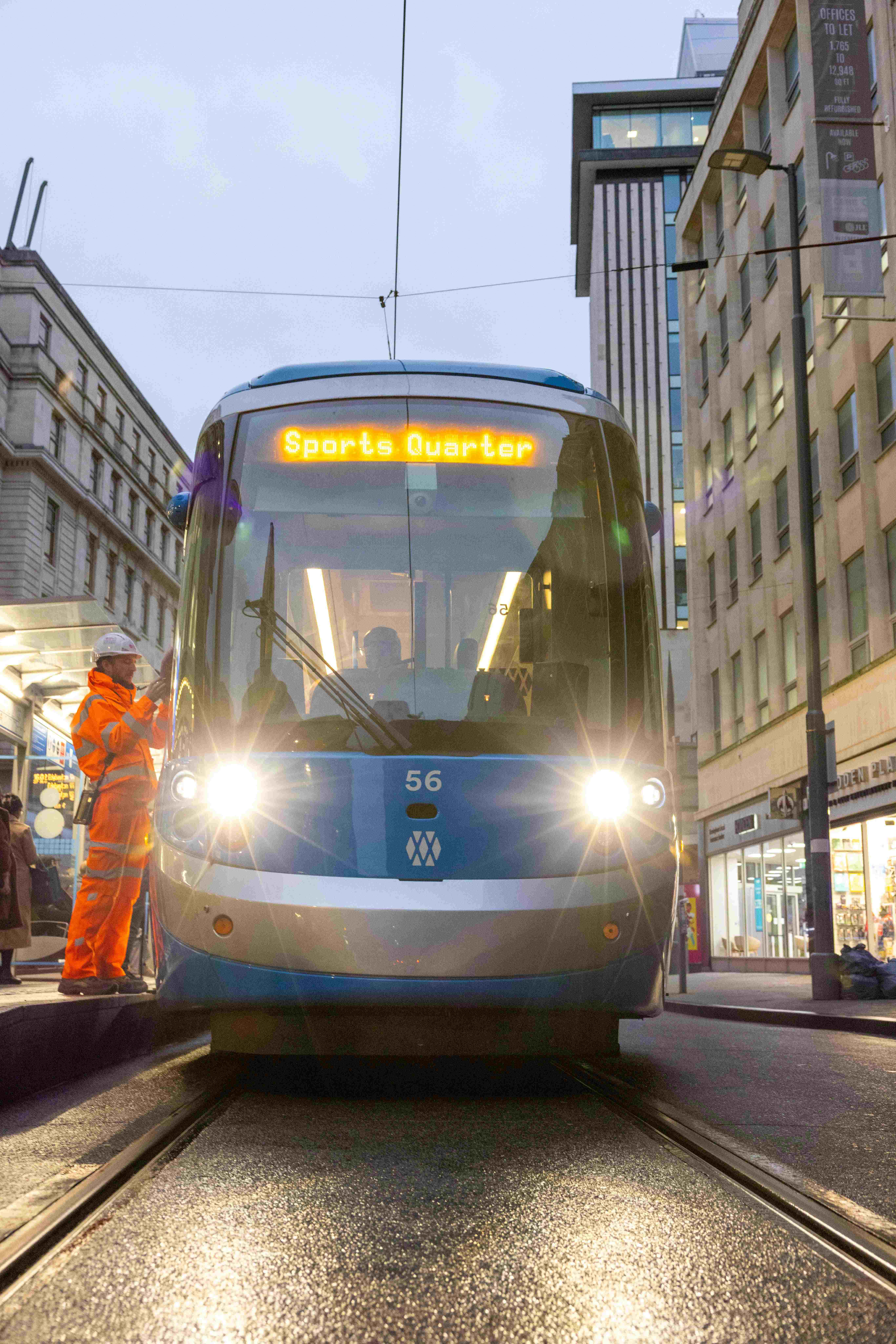 The front of a blue tram on a city street. The headlights are on and the destination board says 'Sports Quarter'.