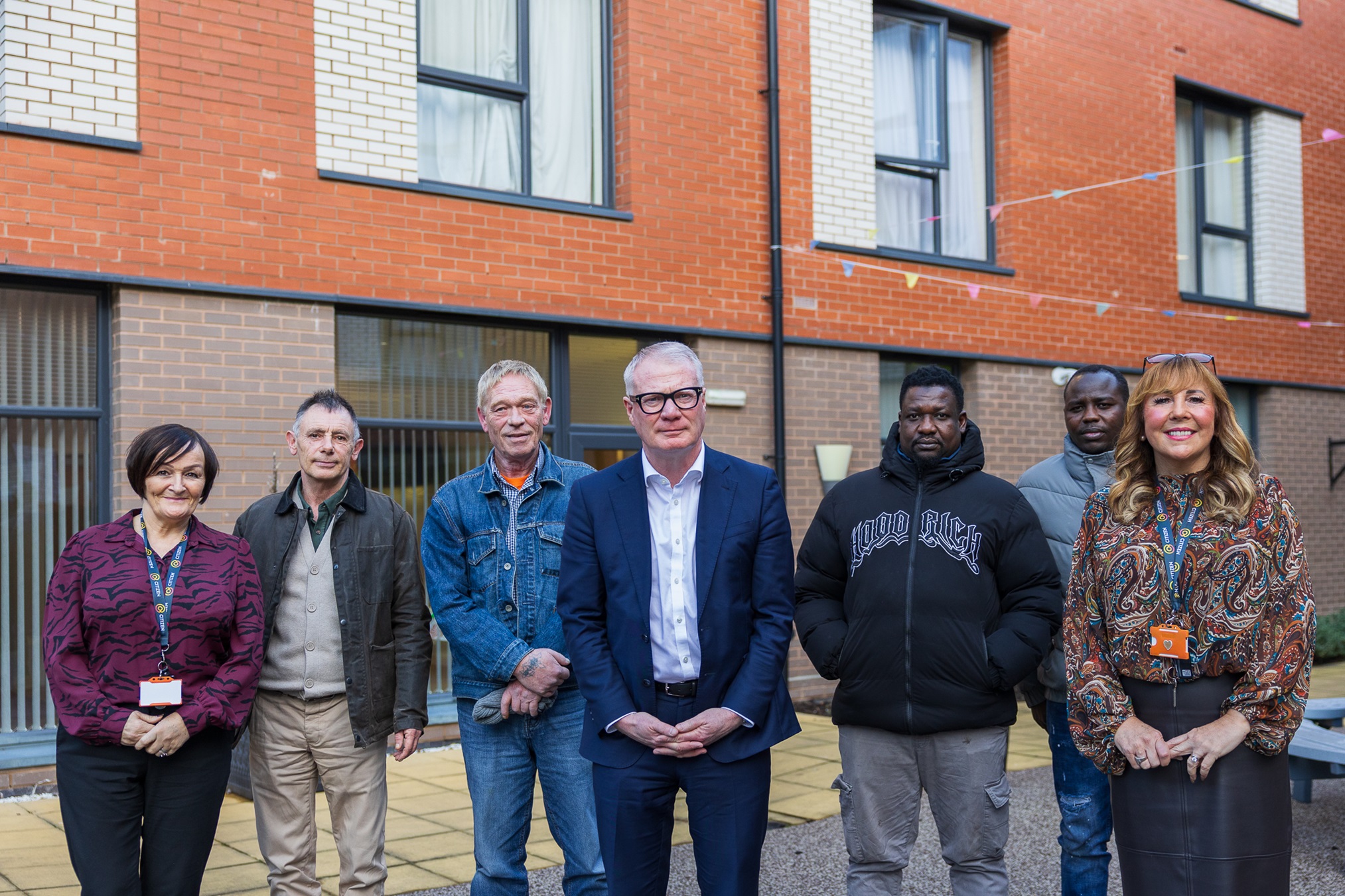 A group of seven diverse people stand smiling outside a modern brick building. They appear friendly and confident, conveying a sense of community and unity.