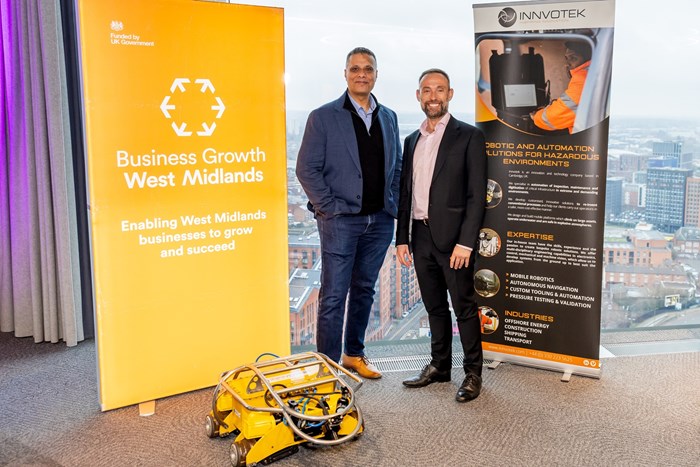Two men in suits stand smiling beside a large orange banner reading "Business Growth West Midlands," with a cityscape seen through the window.