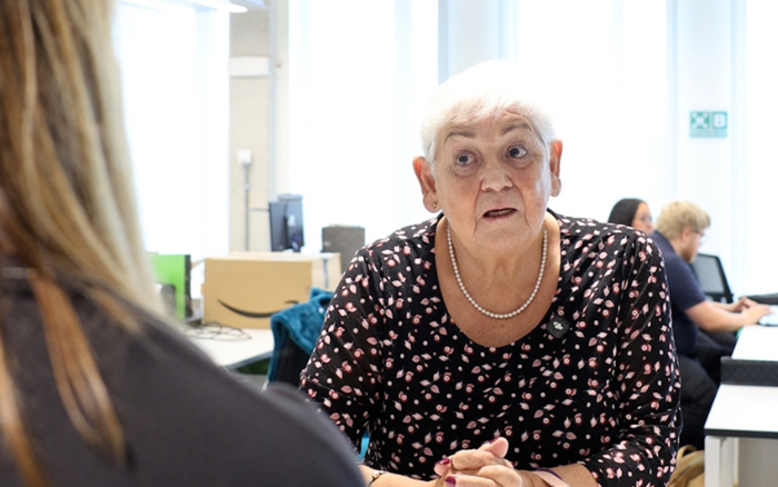 A woman leans forward with hands clasped, speaking to someone across a desk in a bright office; the scene feels attentive and personal.