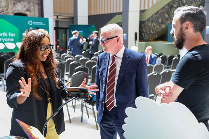A woman in a black blazer energetically gestures while speaking to two men, one in a suit and the other casually dressed, in a conference setting with a banner reading "Growth for Everyone" in the background.