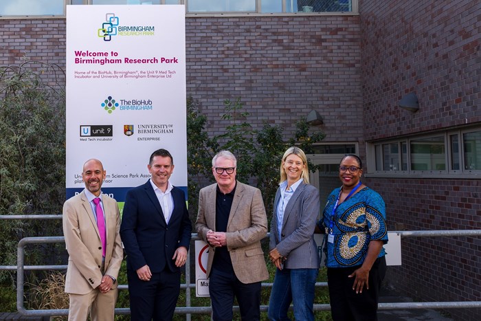 Five people pose outside Birmingham Research Park beside a welcome sign displaying university and health research logos; a professional, welcoming scene.