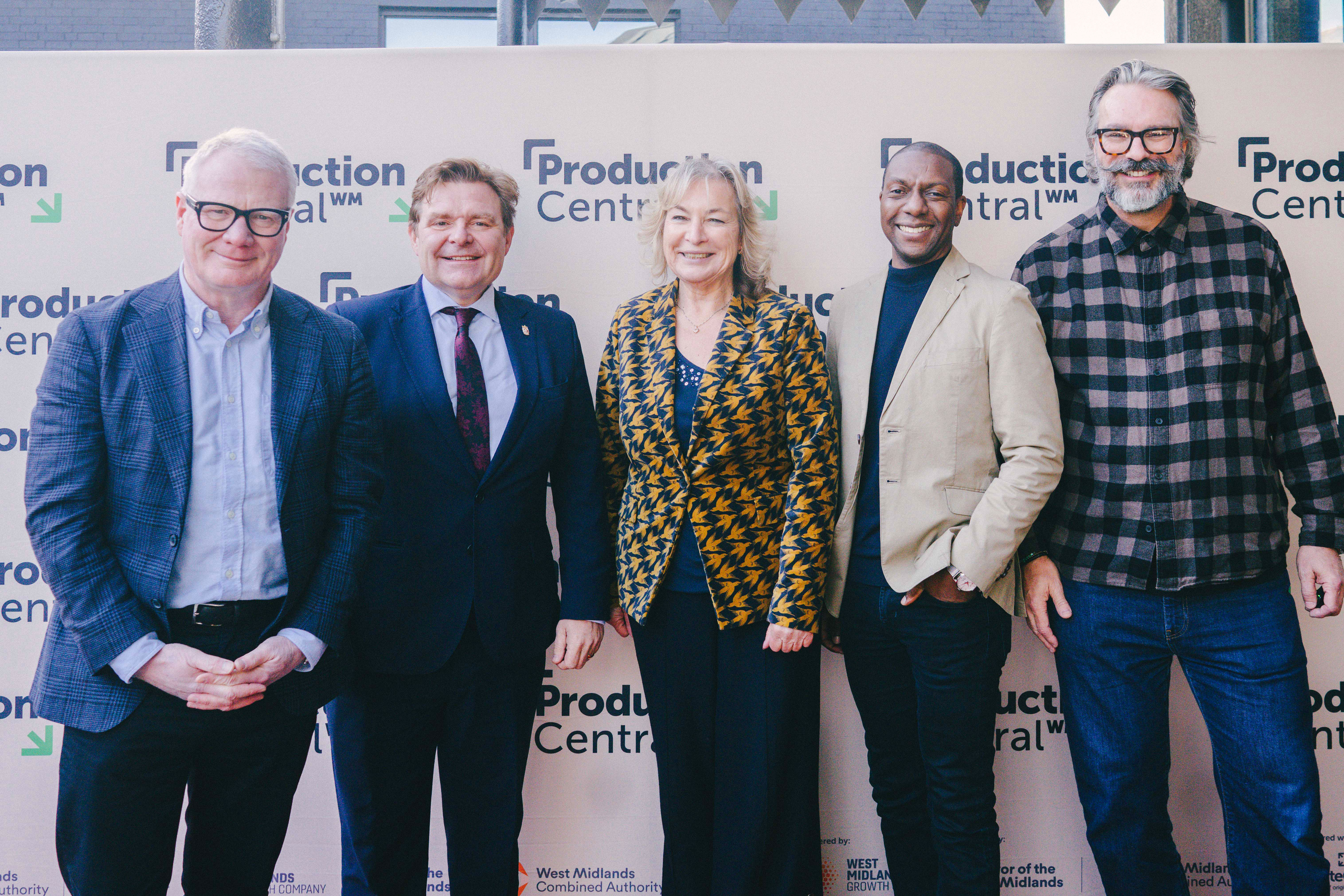 Five adults stand side by side in front of a "Production Central" backdrop, posing for a group photo; all are dressed in business or smart-casual attire.