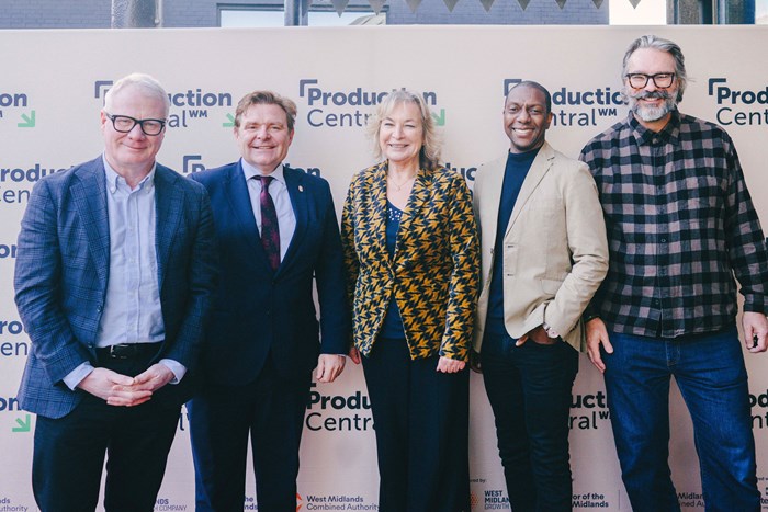 Five adults stand side by side in front of a "Production Central" backdrop, posing for a group photo; all are dressed in business or smart-casual attire.