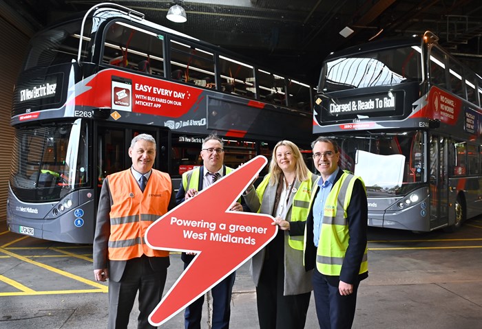 Line up of four officials in hi-vis orange/yellow vests stood in front of two double decker grey buses  in the garage