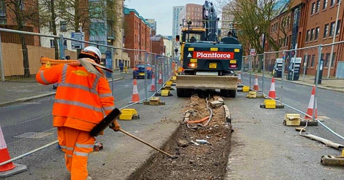 Road worker in orange hi-vis building clothes in foreground and trench dug along city centre road. A digger is further along the trench.