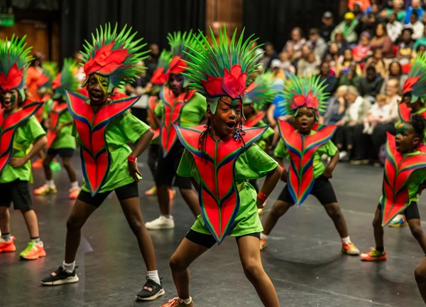 Children performing a dance in bright green costumes with large red and green headpieces, on an indoor stage with an audience seated in the background