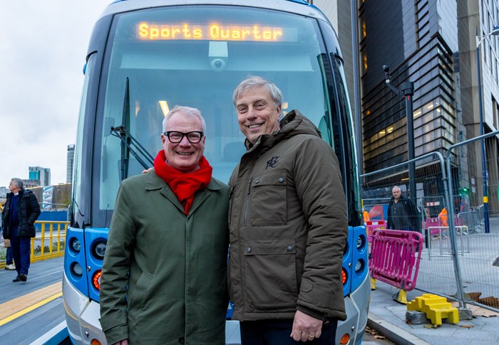 Two men, wrapped up in winter coats, stand in front of a blue tram in a city street. The destination sign on the tram says Sports Quarter