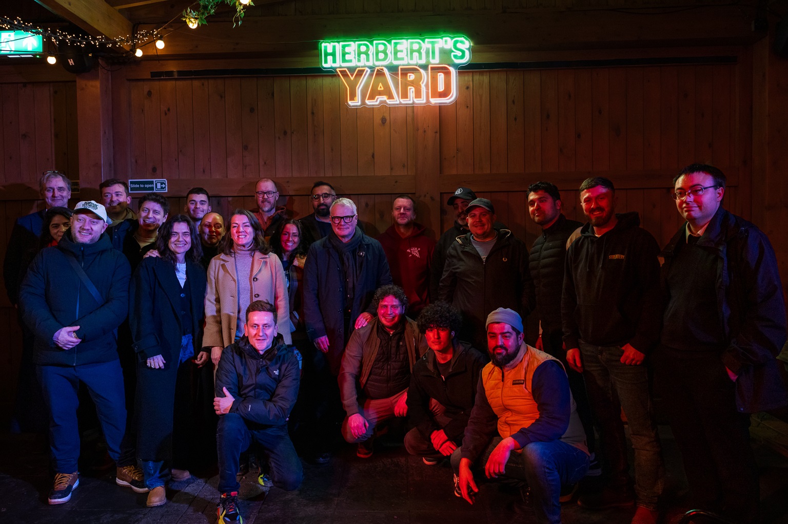 Group photo of 19 people smiling under a neon sign saying "Herbert's Yard." The setting is cozy and warmly lit, suggesting a cheerful gathering.