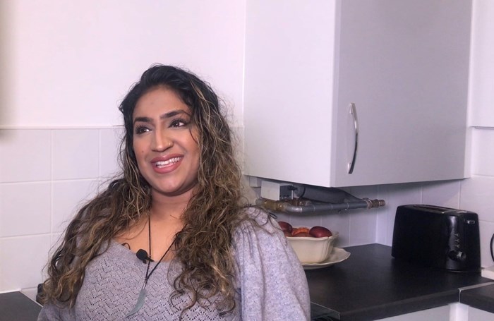 A woman with long curly hair smiles while sitting in a kitchen with white cabinets, a bowl of fruit, and a black toaster on the counter.