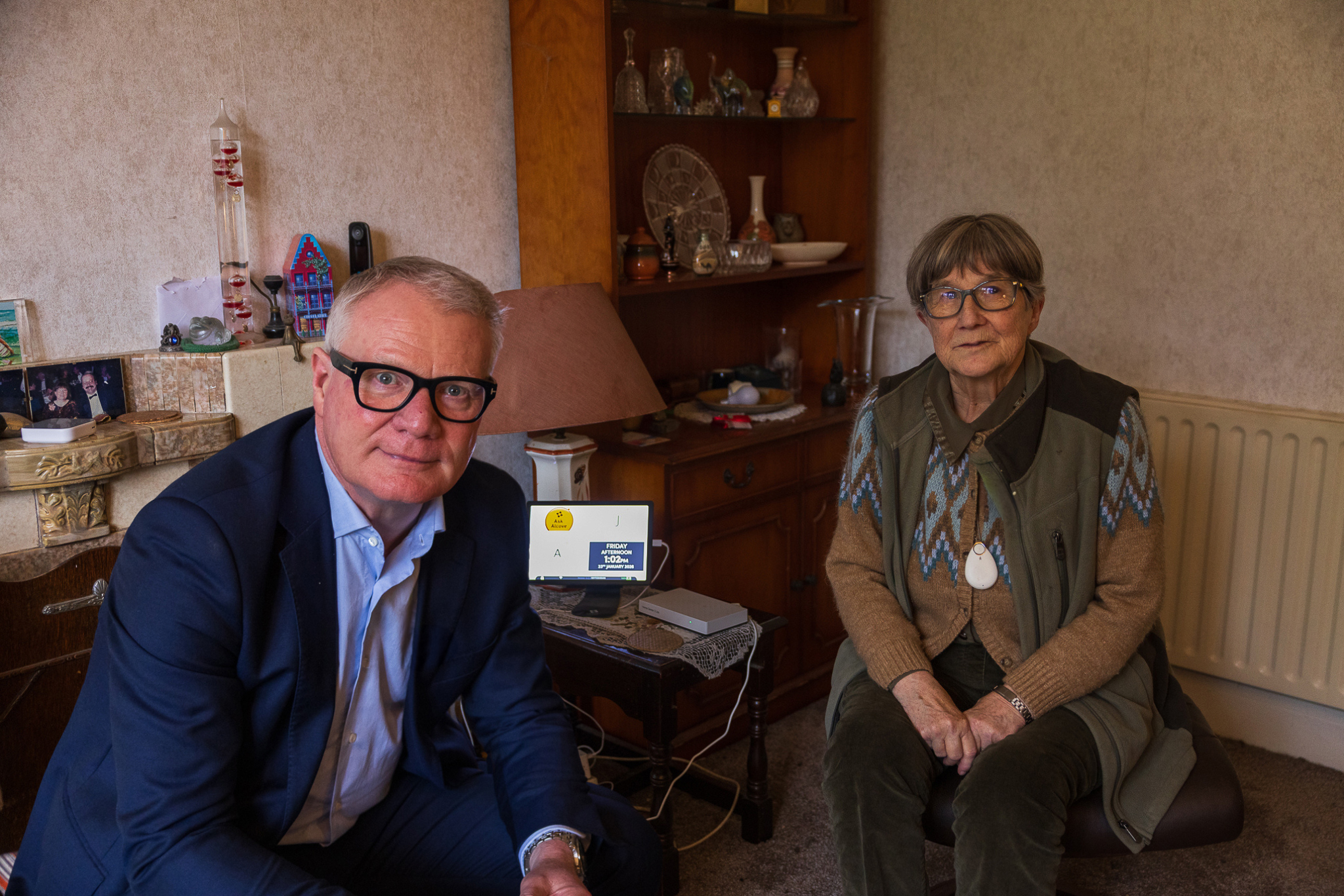 A man in a suit and glasses, and an elderly woman with glasses and a pendant, sit in a cozy living room. Shelves in the background hold various knick-knacks, adding a homely atmosphere.